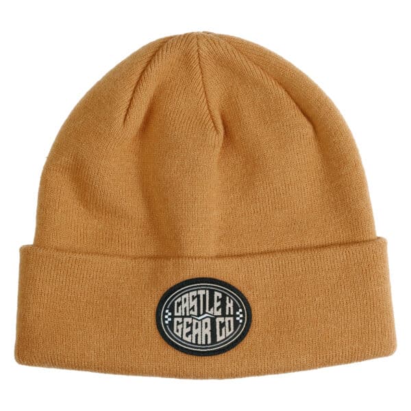 The Live Pom Beanie in mustard yellow features a folded brim and a circular black patch with the white text "CASTLE X GEAR CO" prominently displayed on the front.