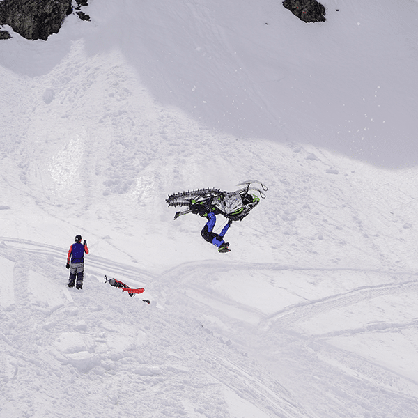 A person performs a backflip on a snowmobile in a snowy mountainous area, with another individual standing nearby, capturing the scene. Tracks are visible in the snow, and the sky is overcast.