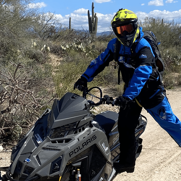 A person wearing a blue and black outfit and a yellow helmet rides a Polaris snowmobile on a dirt path in a desert landscape. There are cacti and shrubs in the background under a clear blue sky.