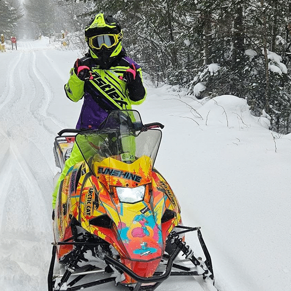 A person in a neon yellow snowmobile suit and helmet sits on a colorful snowmobile in a snowy forest. Their visor is down, and they appear to be showing peace signs with both hands. Snow-covered trees line the background.